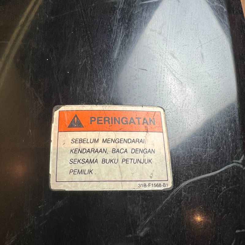 Close-up of the original warning label on the Yamaha XSR155 OEM upper tank cover. Genuine factory fuel tank cover removed from the Neo Muscle project motorcycle, showing the original safety sticker and light cosmetic scratches from normal use.